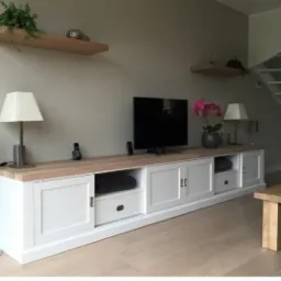 Długa, biała szafka RTV z przesuwnymi drzwiami i drewnianym blatem, ustawiona w salonie z drewnianą podłogą, nad nią wiszące p&oacute;łki i telewizor.