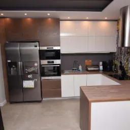 Nowoczesna kuchnia z brązowo-białymi szafkami, dużym srebrnym lod&oacute;wko-zamrażarką, piekarnikiem i kuchenką mikrofalową w zabudowie, oraz blatem z drewnianym wykończeniem i wzorzystą tapetą w tle.