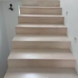 Jasne, drewniane schody wewnętrzne z minimalistycznym oświetleniem punktowym w ścianie, widok z dołu na górę.