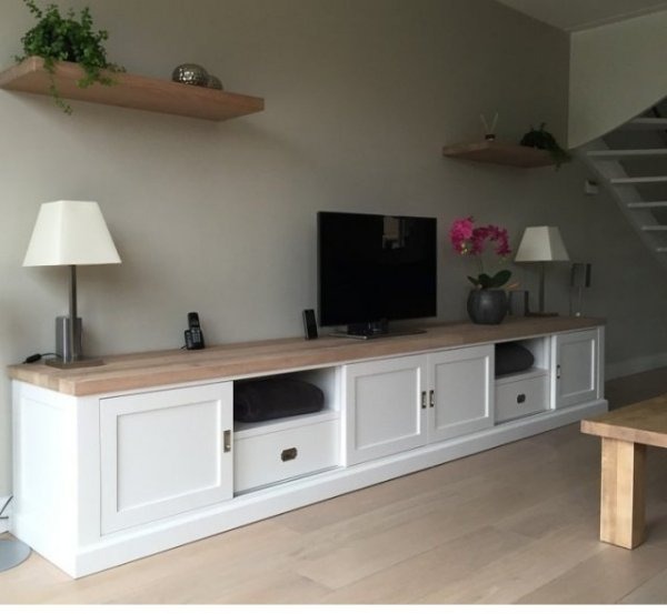 Długa, biała szafka RTV z przesuwnymi drzwiami i drewnianym blatem, ustawiona w salonie z drewnianą podłogą, nad nią wiszące półki i telewizor.