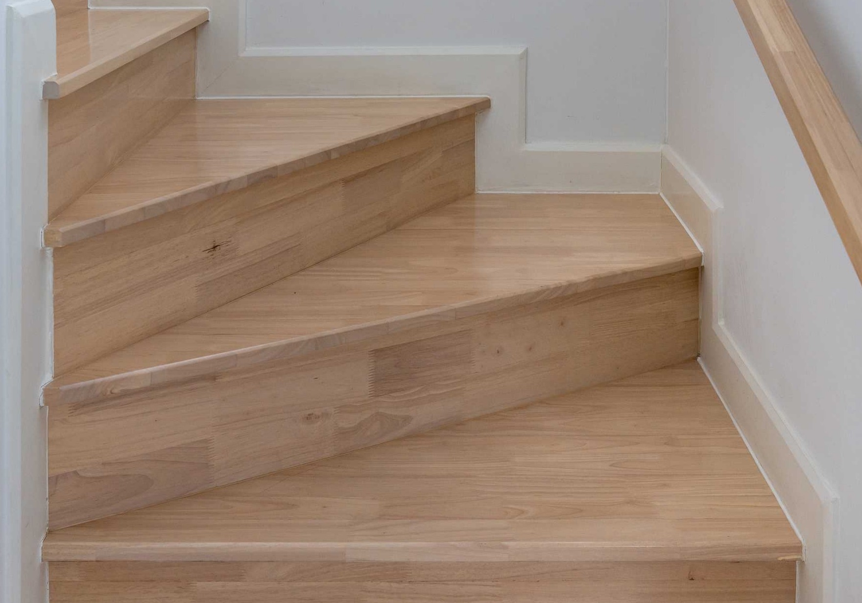 Jasne, drewniane stopnie schodów z widoczną strukturą drewna, białe listwy przypodłogowe i fragment poręczy.