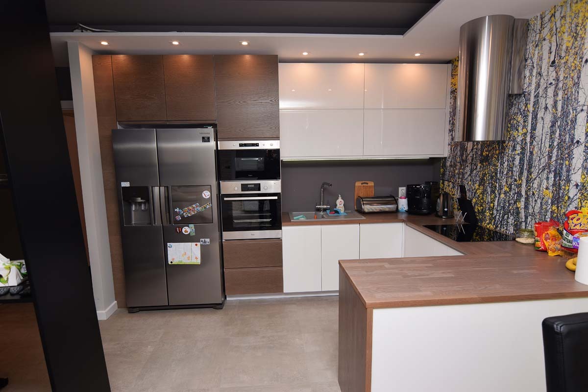 Nowoczesna kuchnia z brązowo-białymi szafkami, dużym srebrnym lodówko-zamrażarką, piekarnikiem i kuchenką mikrofalową w zabudowie, oraz blatem z drewnianym wykończeniem i wzorzystą tapetą w tle.