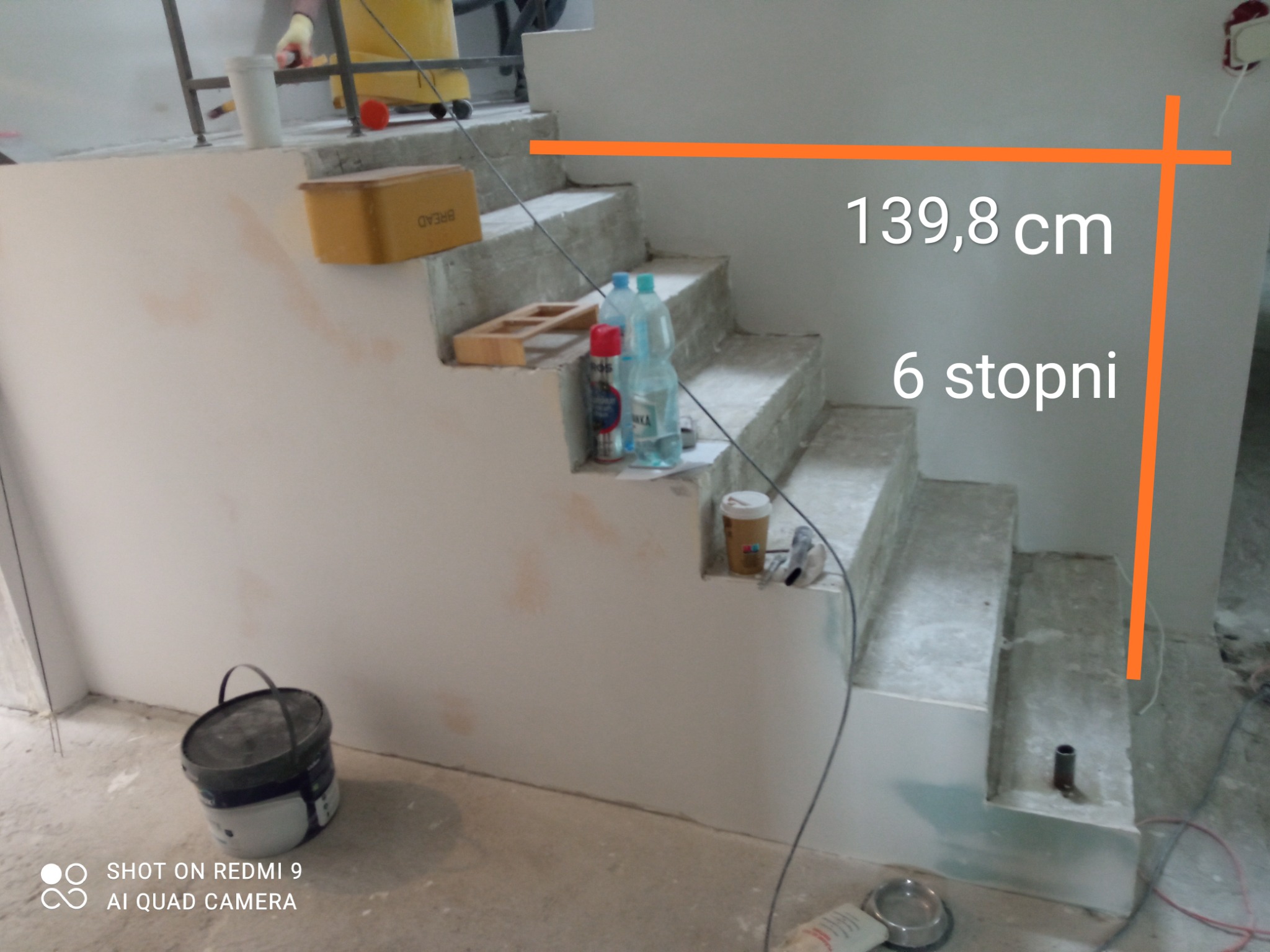 Surowe, betonowe schody z sześcioma stopniami w trakcie budowy, z naniesionymi pomiarami 139,8 cm, widoczne materiały budowlane i wiadro z farbą.