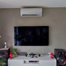 Biały klimatyzator zamontowany nad czarnym telewizorem w salonie z beżową ścianą, pod nim biała szafka RTV z dekoracjami i głośnikami Manta po bokach.