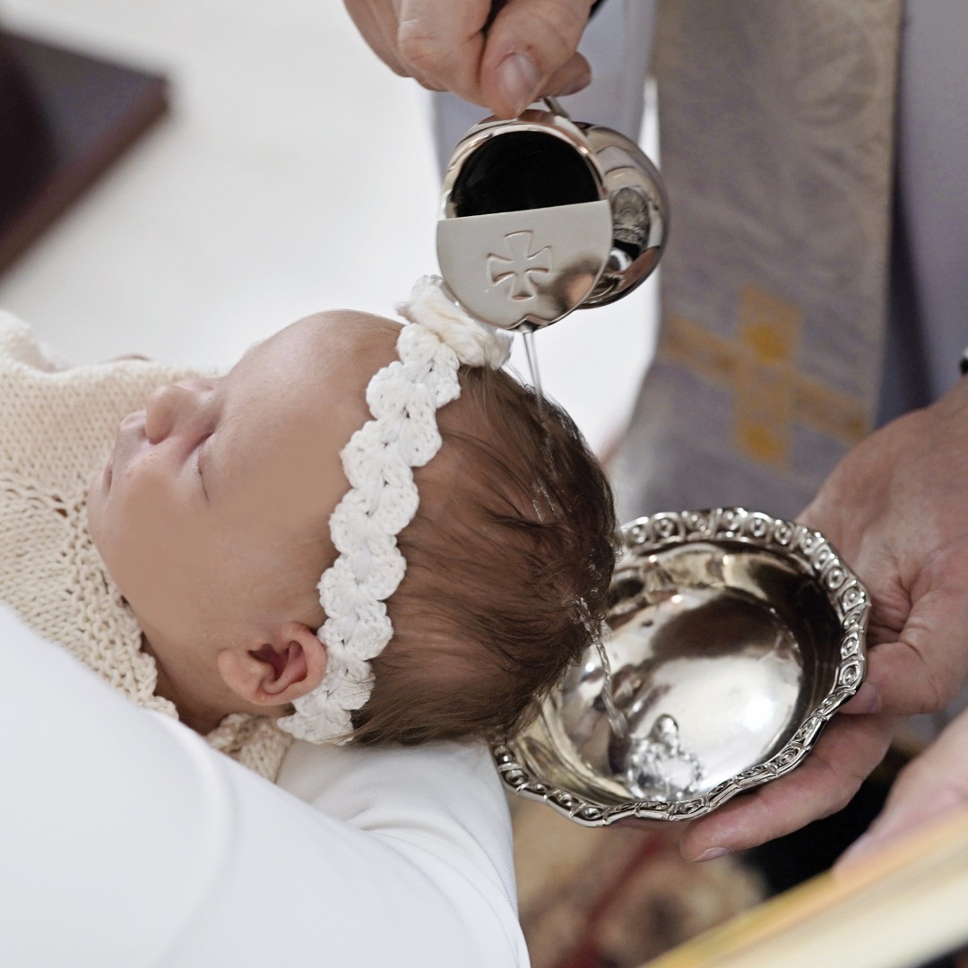 Niemowlę w białej opasce podczas chrztu, woda polewana na głowę z ozdobnego naczynia przez ręce osoby dorosłej.