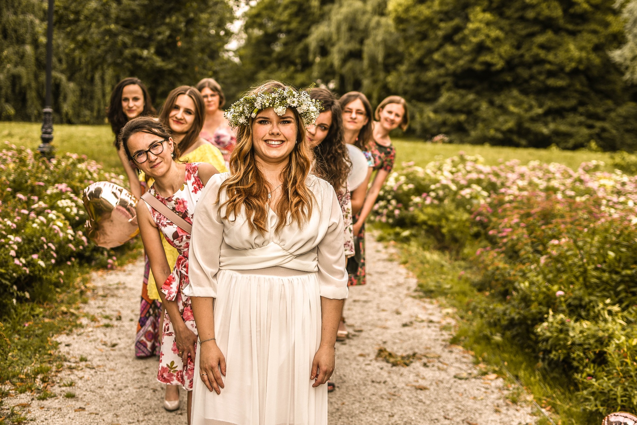 Przyszła panna młoda w białej sukni i wianku na głowie, otoczona przez druhny w kwiecistych sukienkach, pozuje na ścieżce w parku, trzymając balon w kształcie serca.