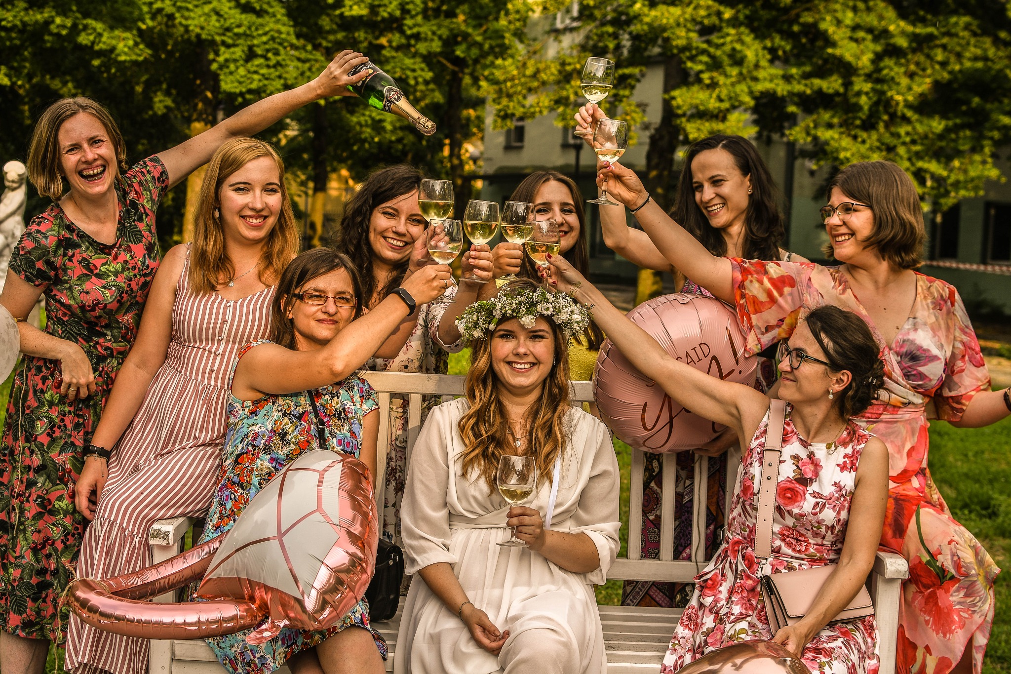 Grupa kobiet świętuje na świeżym powietrzu, wznosząc toast winem; panna młoda w białej sukience i wianku siedzi na ławce, otoczona druhnami z balonami w kształcie pierścionka i napisem 'She said yes'.