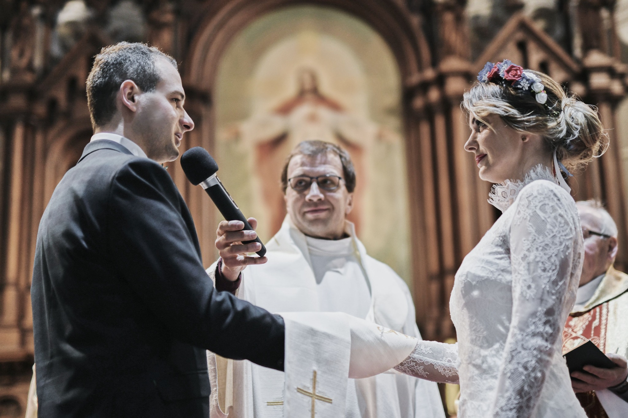 Para młoda podczas ceremonii ślubnej w kościele, panna młoda w koronkowej sukni i wianku, pan młody z mikrofonem, w tle ksiądz.