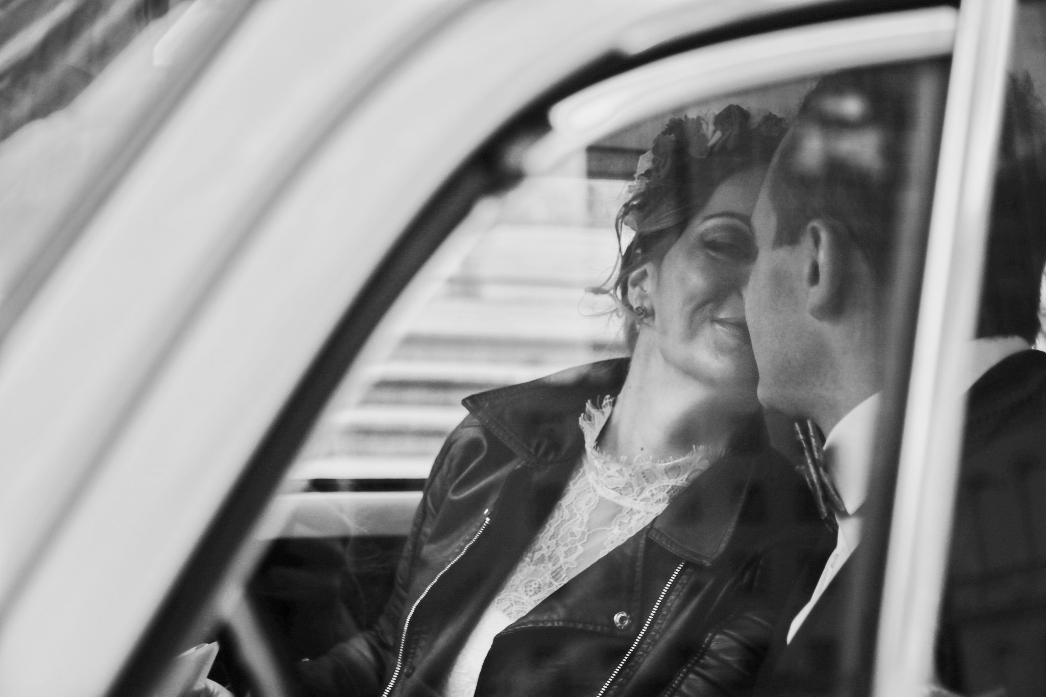 Czarno-białe zdjęcie pary młodej w samochodzie, panna młoda w skórzanej kurtce i koronkowej sukience, pan młody w garniturze, widoczni przez szybę.