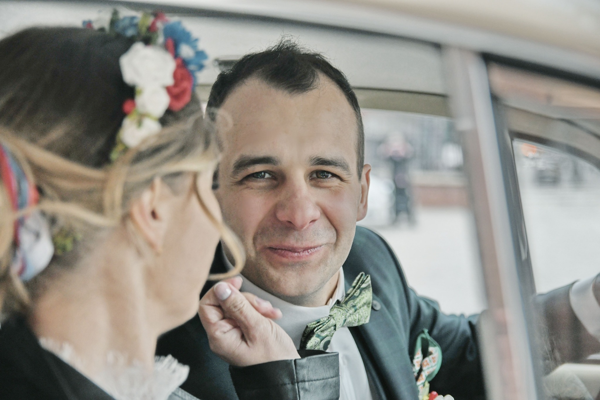 Uśmiechnięty pan młody w zielonej muszce i butonierce, trzymający dłoń panny młodej w samochodzie, z widocznym kwiatowym wiankiem na jej głowie.