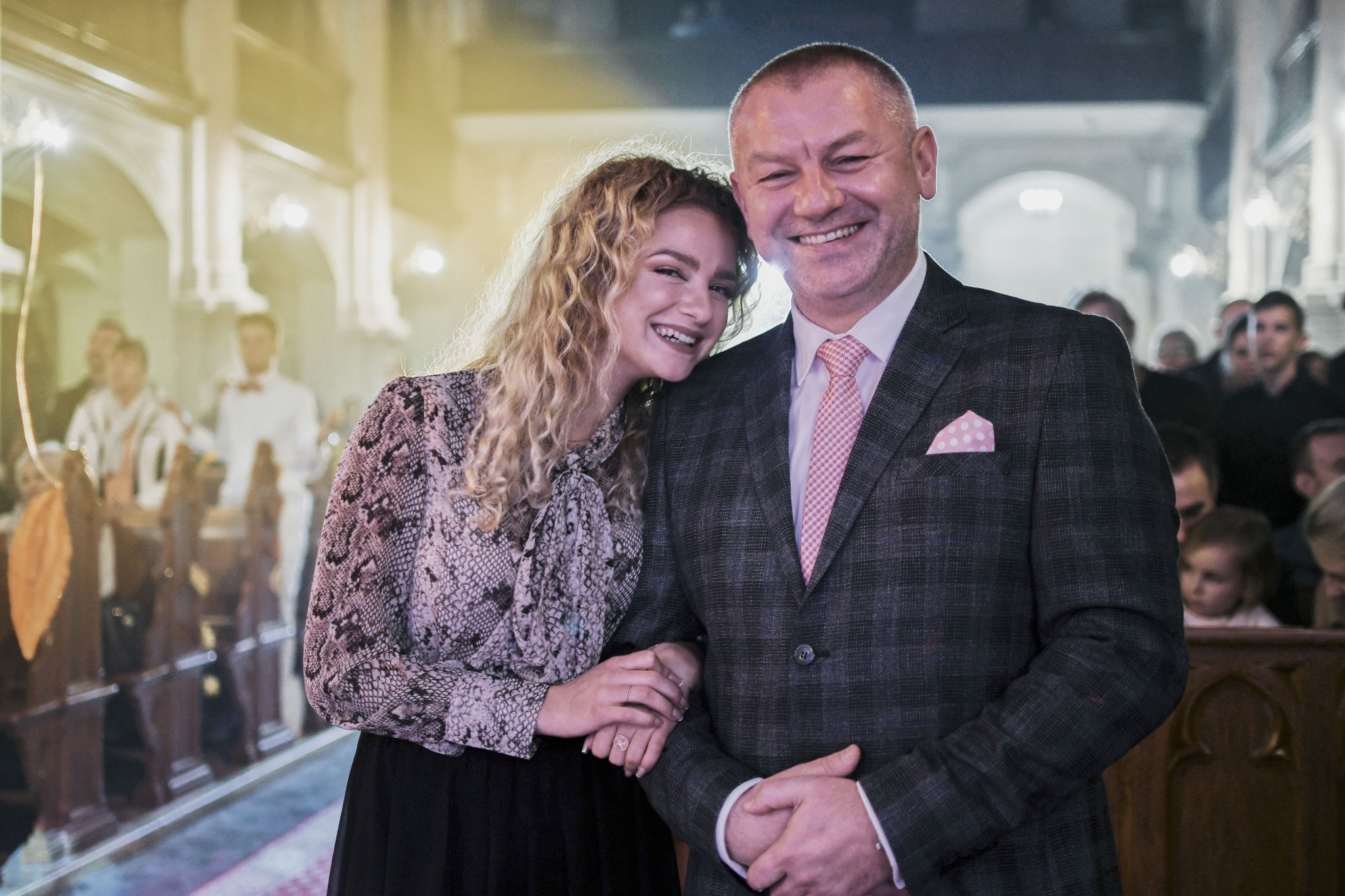 Uśmiechnięta para: młoda kobieta z kręconymi włosami opiera głowę na ramieniu eleganckiego mężczyzny w garniturze z różową poszetką w kropki, w tle kościół i goście.