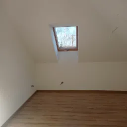 Poddasze po remoncie: świeżo pomalowane białe ściany, drewnopodobne panele podłogowe, widoczne okno dachowe z widokiem na drzewa.