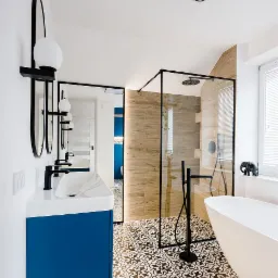 Nowoczesna łazienka w Płocku z niebieską szafką pod umywalką, kabiną prysznicową typu walk-in z drewnopodobnymi płytkami oraz wolnostojącą wanną z czarną armaturą i dekoracyjną figurką słonia...