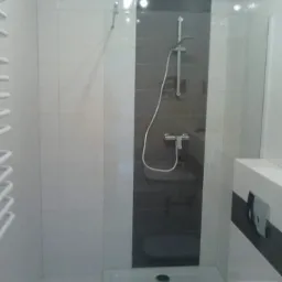 Nowoczesna kabina prysznicowa z szarą ścianką i białymi płytkami, widoczny brodzik, bateria i słuchawka prysznicowa oraz fragment umywalki z baterią.