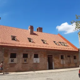 Ceglany budynek z czerwoną dach&oacute;wką i otwartymi oknami dachowymi, widoczne dwa kominy, ściany z ubytkami tynku i zaklejonymi oknami, chodnik z kostki brukowej, błękitne niebo.