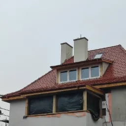 Dom w trakcie budowy/remontu: widoczny nowy dach z czerwoną dachówką, okna dachowe, kominy, okna na poddaszu z folią ochronną, rusztowania i ocieplenie ścian zewnętrznych.