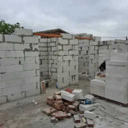 Wznoszenie ścian działowych z białych bloczków na budowie, widoczne spoiny z zaprawy, cegły, narzędzie do cięcia i worki z zaprawą, pochmurne niebo w tle.