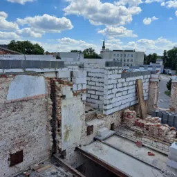 Widok z góry na budowę z częściowo zburzoną ceglaną ścianą, nowymi bloczkami betonowymi i elementami konstrukcyjnymi stalowymi, z widocznym niebieskim niebem w tle.