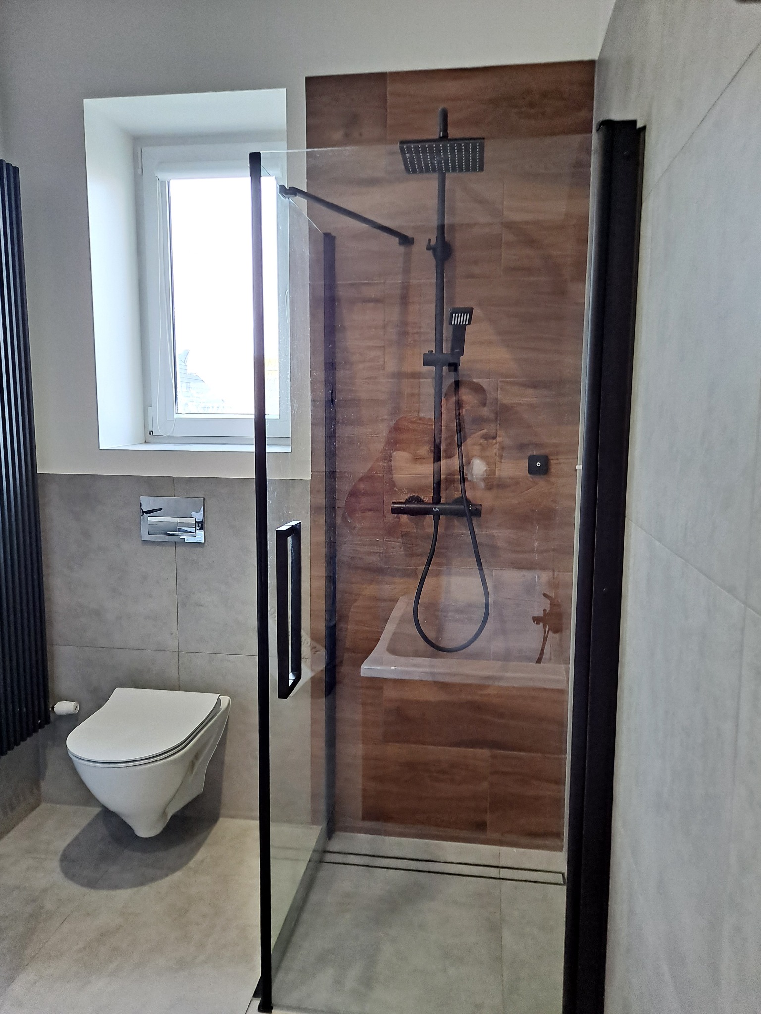Nowoczesna łazienka z kabiną prysznicową typu walk-in, wykończona płytkami imitującymi drewno i szarością, z czarną armaturą i grzejnikiem drabinkowym.