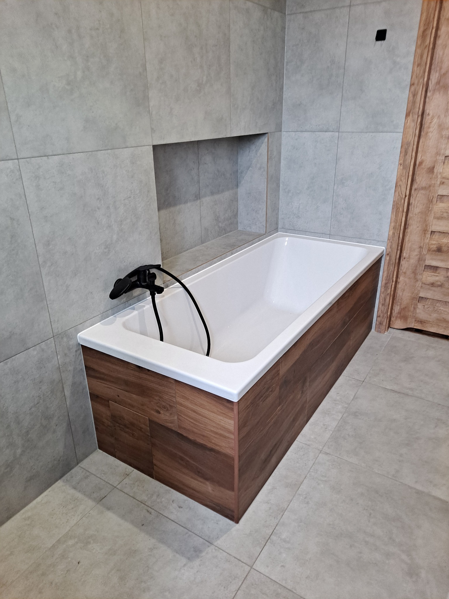 Nowoczesna łazienka z wanną obłożoną panelami drewnopodobnymi, czarna bateria, szare płytki ścienne i podłogowe oraz drewniane drzwi w tle.