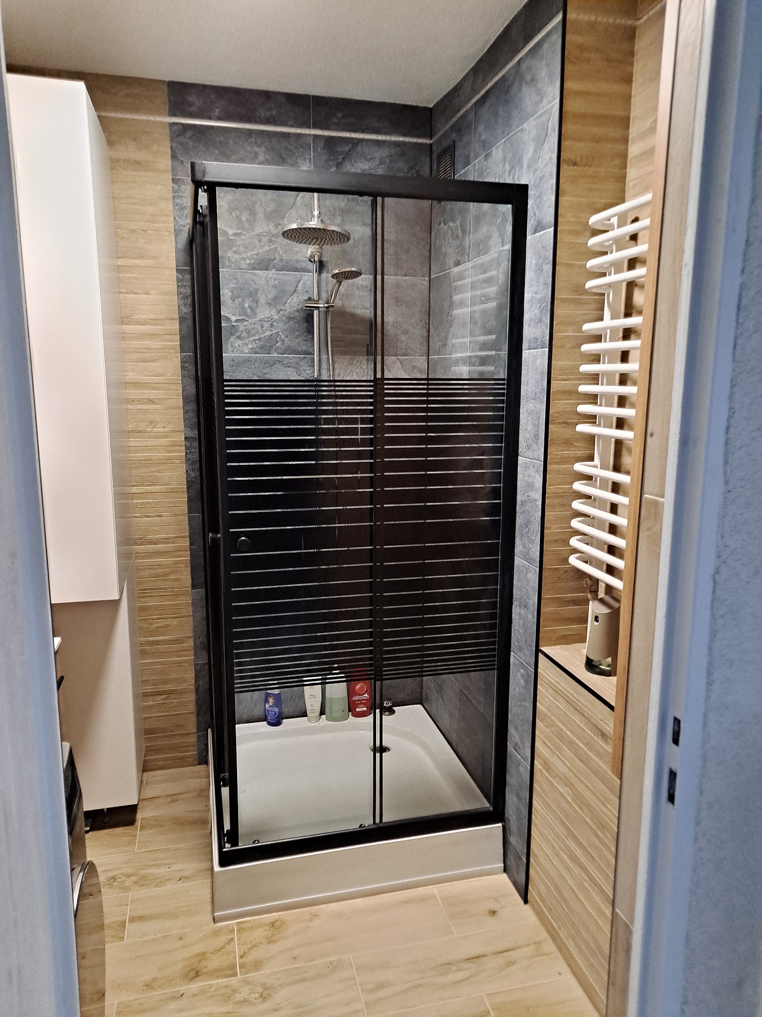 Nowoczesna kabina prysznicowa z czarną ramą i deszczownicą w wykończonej łazience z szarymi i beżowymi płytkami, widoczne kosmetyki wewnątrz kabiny.