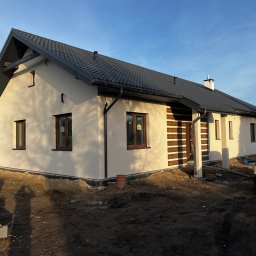 ART-REM - Otynkowany dom jednorodzinny z ciemną dachówką i brązowymi oknami, widoczny wczesnym rankiem, z niezagospodarowanym terenem wokół, przed domem zaparkowany czarny samochód.