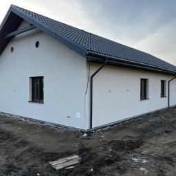 ART-REM - Nowo otynkowany dom jednorodzinny z ciemną dachówką i brązowymi oknami, widok z zewnątrz na plac budowy z ziemią i widocznymi elementami instalacji elektrycznej.