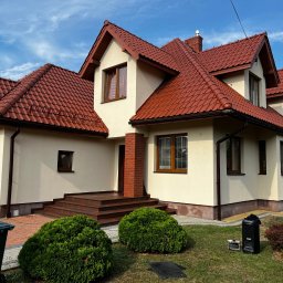 ART-REM - Dwukondygnacyjny dom z czerwonym dachem z dachówki ceramicznej i beżową elewacją, mężczyzna na drabinie montuje oświetlenie zewnętrzne, widoczne krzewy i kosz na śmieci.