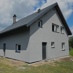 ART-REM - Szary dom jednorodzinny z ciemnoszarą dachówką i tynkiem strukturalnym, widoczny z boku w słoneczny dzień, z elementami wykończeniowymi wokół budynku i rozłożonymi wężami ogrodowymi.