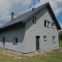 Szary dom jednorodzinny z ciemnoszarą dachówką i tynkiem strukturalnym, widoczny z boku w słoneczny dzień, z elementami wykończeniowymi wokół budynku i rozłożonymi wężami ogrodowymi.