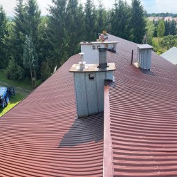 ART-REM - Czerwony, falisty dach z blachy trapezowej, z widocznymi kominami i pracownikiem w oddali, na tle zieleni drzew i zabudowań.