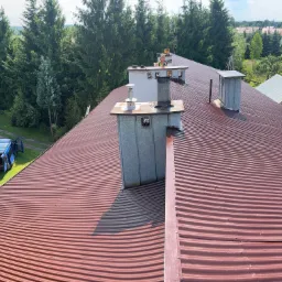 Czerwony, falisty dach z blachy trapezowej, z widocznymi kominami i pracownikiem w oddali, na tle zieleni drzew i zabudowań.