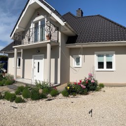 ART-REM - Jasnoszary dom jednorodzinny z balkonem i czarnym dachem, otoczony kwitnącymi różami w ogrodzie i wysypany żwirem.