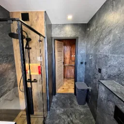 Nowoczesna łazienka z kabiną prysznicową typu walk-in z czarną ramą, szarymi płytkami imitującymi marmur na ścianach i podłodze, oraz drewnianymi akcentami wewnątrz kabiny i w sąsiednim...
