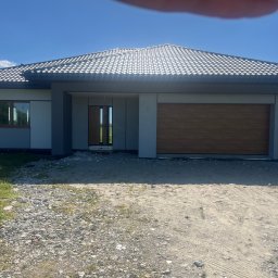 ART-REM - Nowoczesny dom jednorodzinny z szarą elewacją i drewnianą bramą garażową w słoneczny dzień. Widoczny fragment drabiny z pracownikiem.