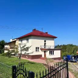 Dwupiętrowy dom z czerwonym dachem i beżową elewacją, widoczny zza czarnego, ozdobnego ogrodzenia. Na podjeździe stoi niebieski bus z otwartymi drzwiami.