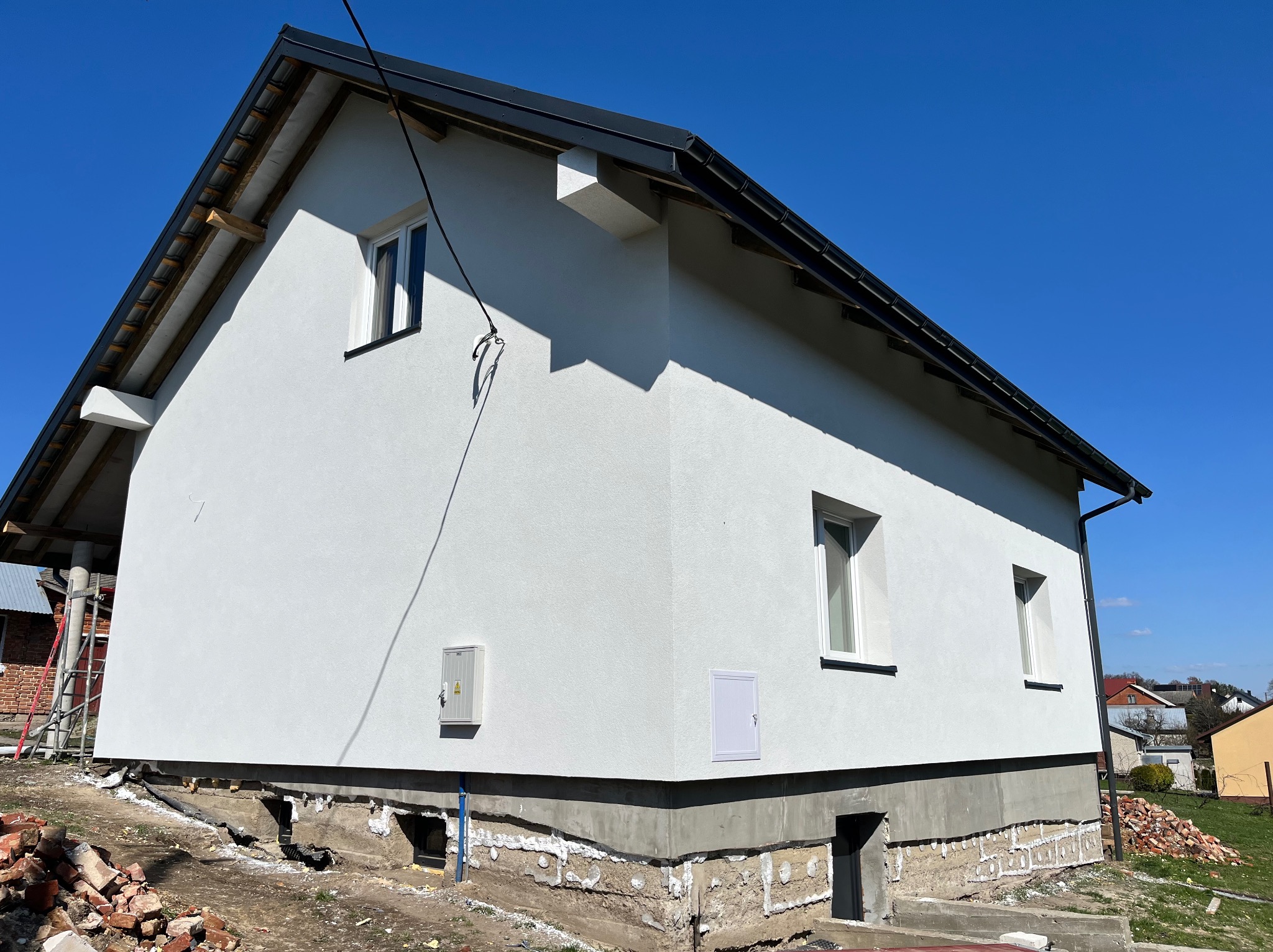 Świeżo otynkowana, biała elewacja domu jednorodzinnego z widocznymi fragmentami fundamentów podczas prac wykończeniowych, na tle błękitnego nieba.