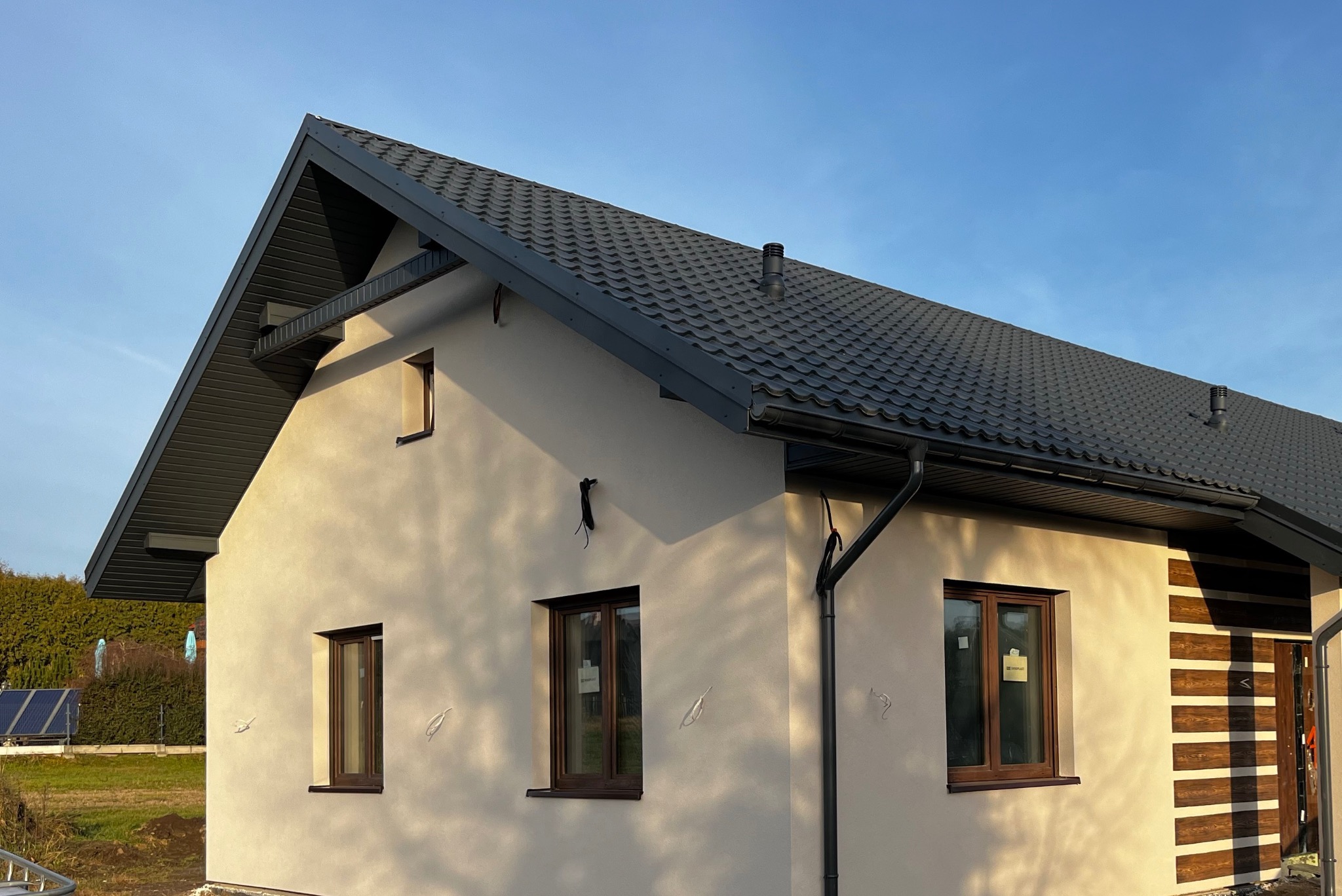 Nowoczesny dom jednorodzinny z jasną elewacją, ciemnym dachem z blachodachówki i brązowymi oknami, widok z zewnątrz w słoneczny dzień, z widocznymi elementami instalacji na elewacji.