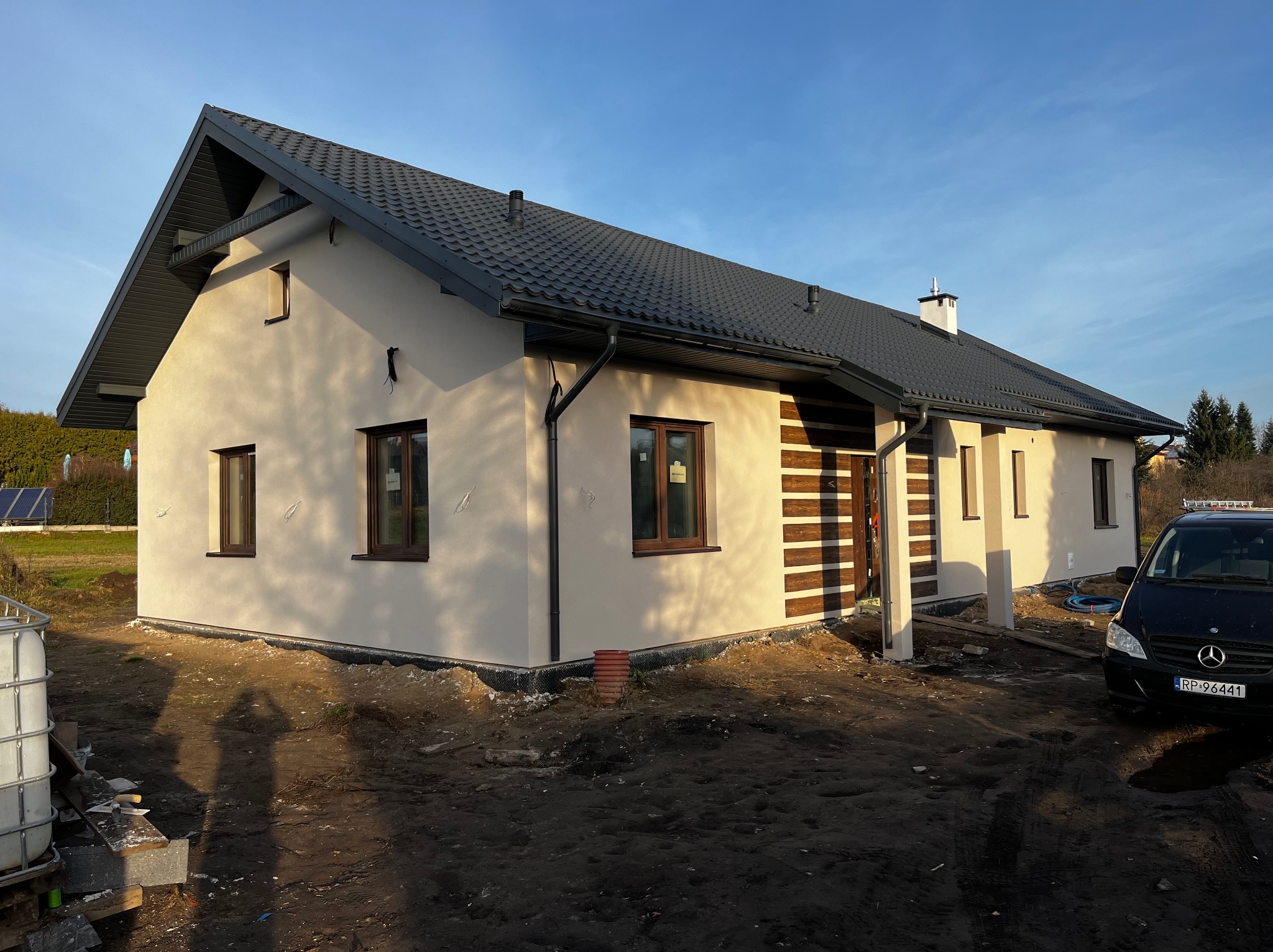 Otynkowany dom jednorodzinny z ciemną dachówką i brązowymi oknami, widoczny wczesnym rankiem, z niezagospodarowanym terenem wokół, przed domem zaparkowany czarny samochód.