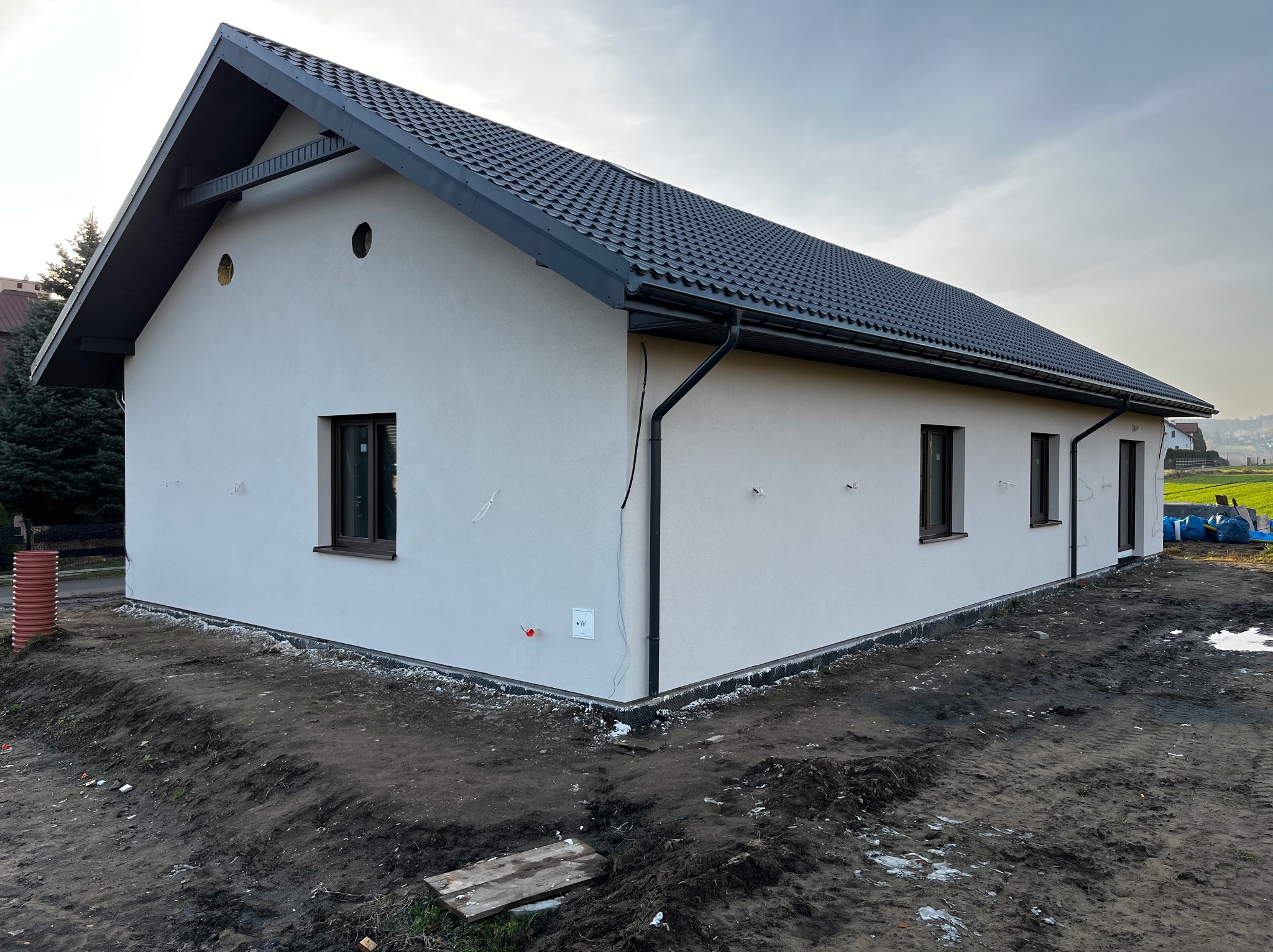 Nowo otynkowany dom jednorodzinny z ciemną dachówką i brązowymi oknami, widok z zewnątrz na plac budowy z ziemią i widocznymi elementami instalacji elektrycznej.