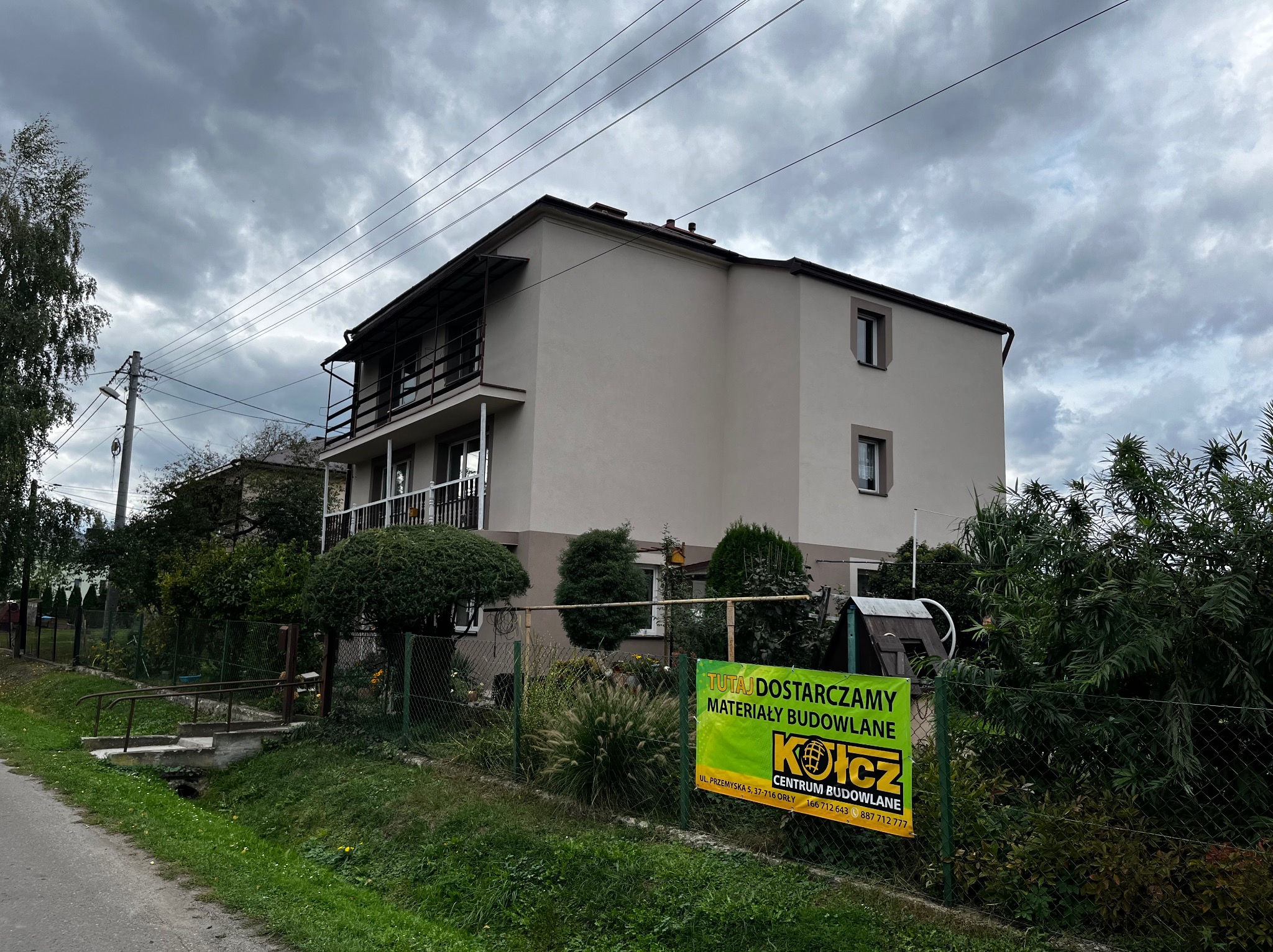 Dwupiętrowy dom z balkonem, świeżo po pracach elewacyjnych, widoczny zza ogrodzenia z reklamą centrum budowlanego.