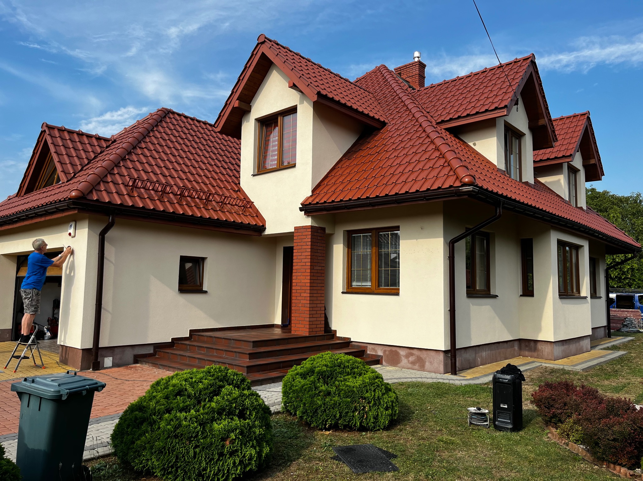 Dwukondygnacyjny dom z czerwonym dachem z dachówki ceramicznej i beżową elewacją, mężczyzna na drabinie montuje oświetlenie zewnętrzne, widoczne krzewy i kosz na śmieci.