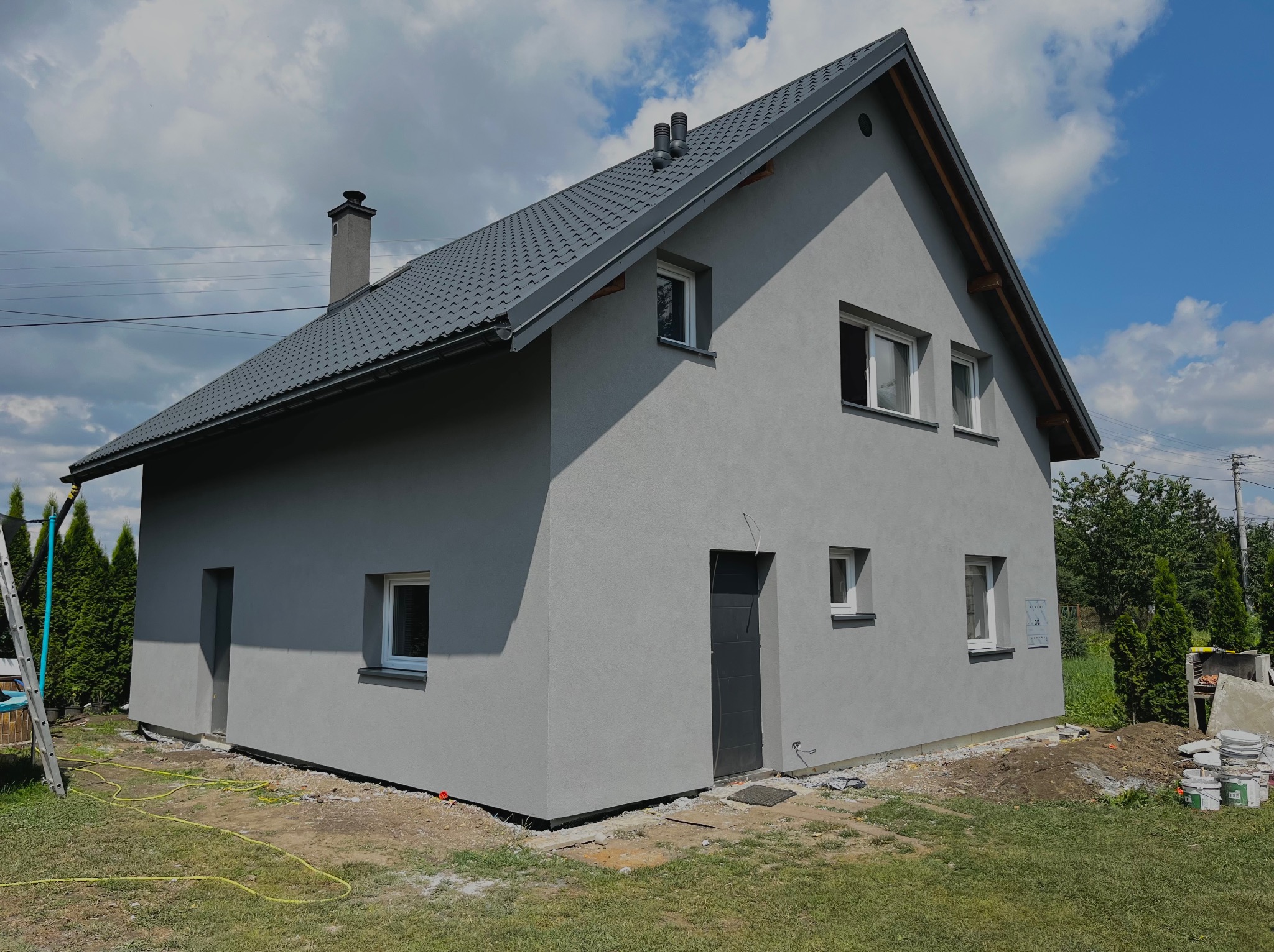 Szary dom jednorodzinny z ciemnoszarą dachówką i tynkiem strukturalnym, widoczny z boku w słoneczny dzień, z elementami wykończeniowymi wokół budynku i rozłożonymi wężami ogrodowymi.