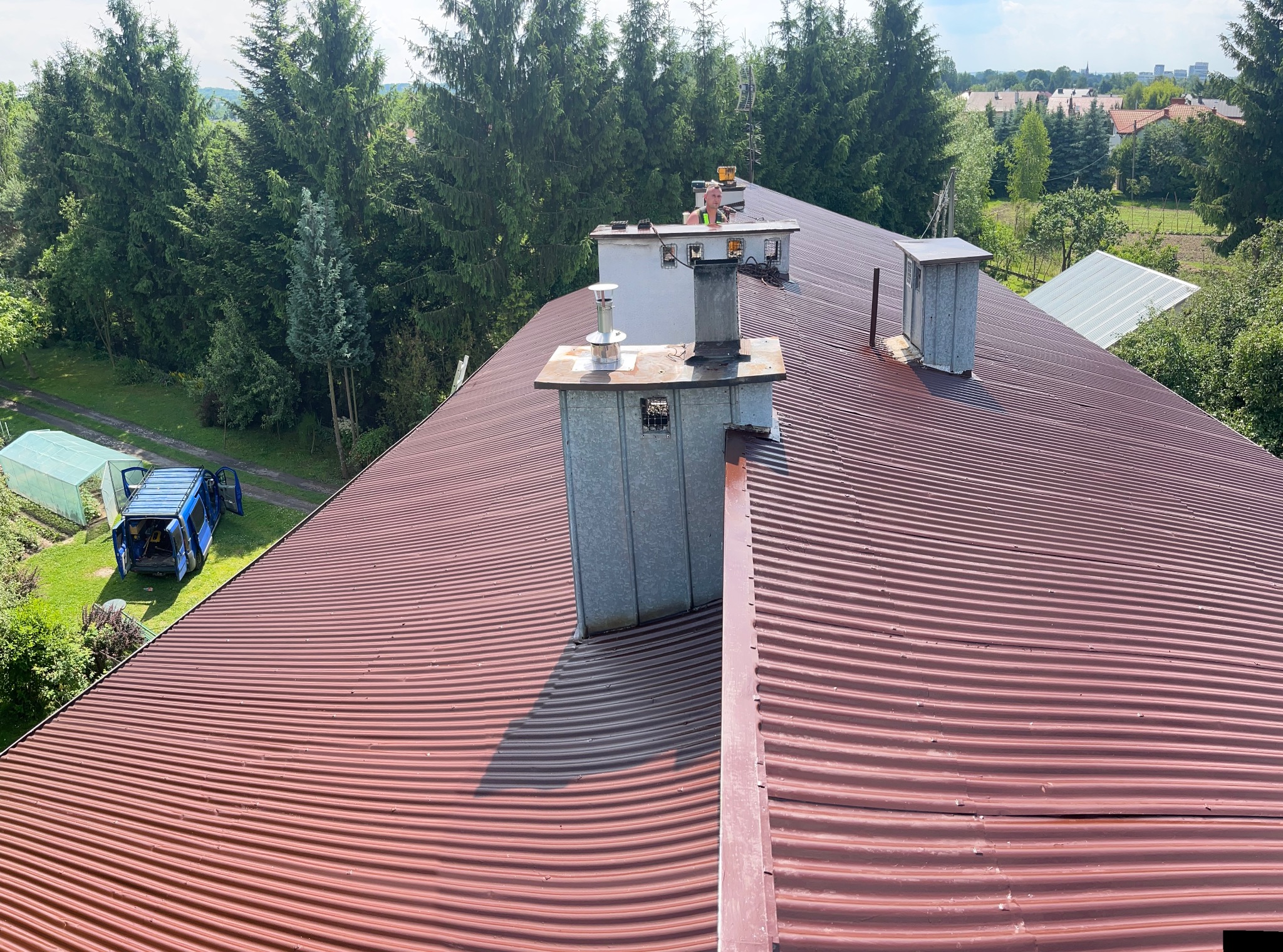 Czerwony, falisty dach z blachy trapezowej, z widocznymi kominami i pracownikiem w oddali, na tle zieleni drzew i zabudowań.