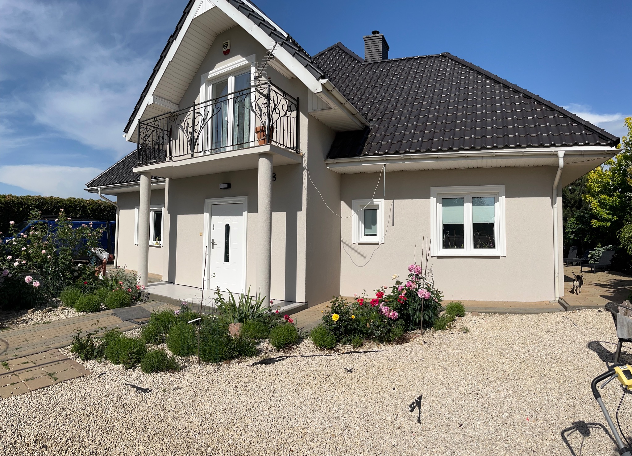 Jasnoszary dom jednorodzinny z balkonem i czarnym dachem, otoczony kwitnącymi różami w ogrodzie i wysypany żwirem.