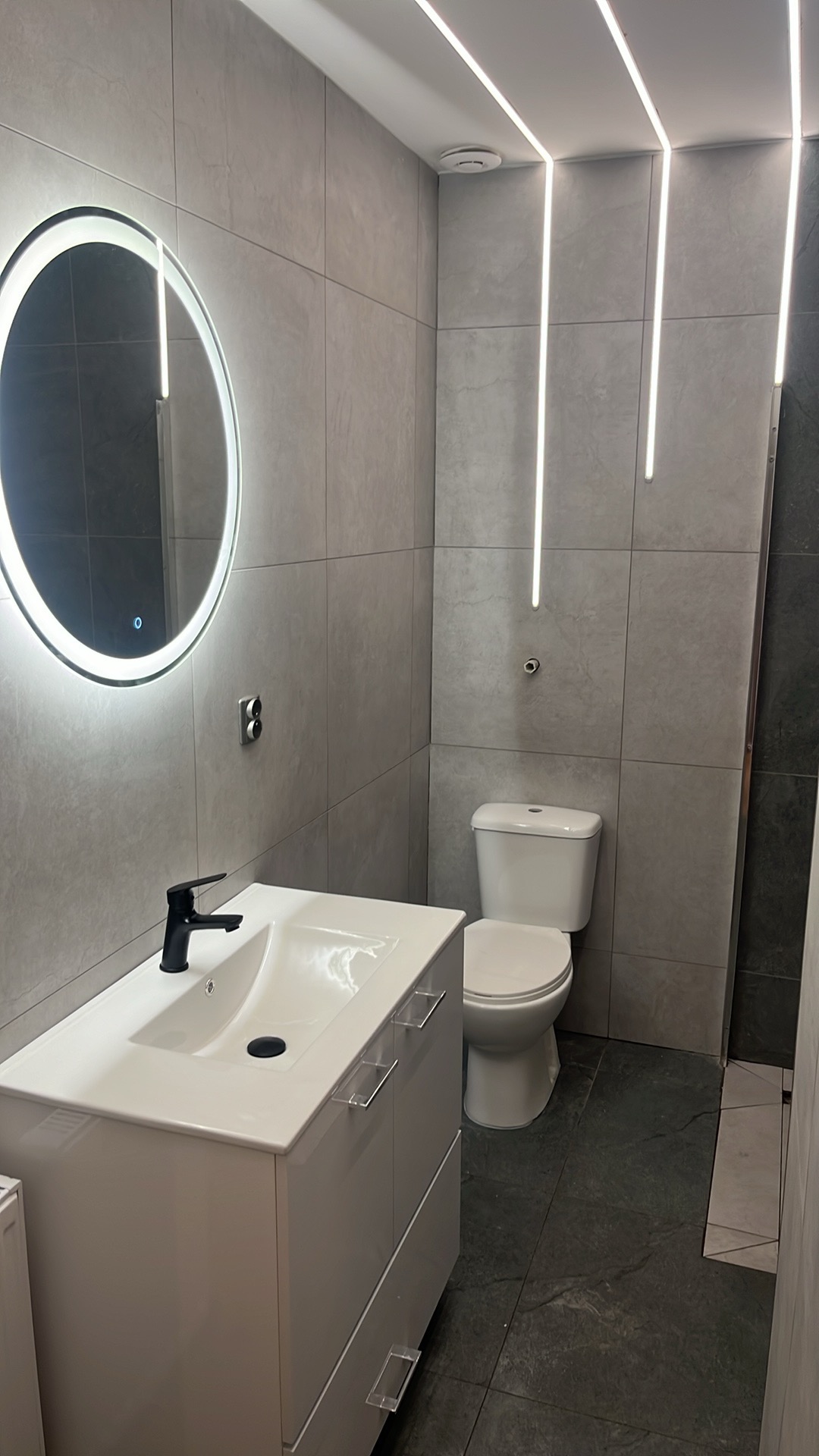 Nowoczesna łazienka z szarymi płytkami, podświetlanym lustrem i liniowym oświetleniem LED na suficie, białą umywalką z czarną armaturą i toaletą.