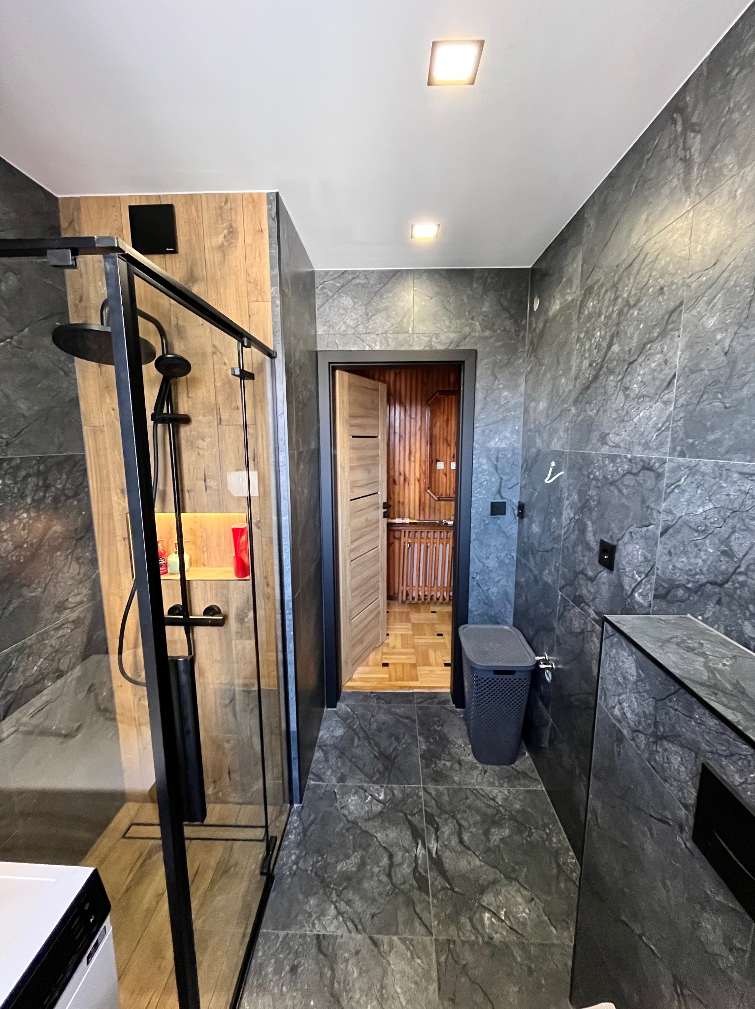 Nowoczesna łazienka z kabiną prysznicową typu walk-in z czarną ramą, szarymi płytkami imitującymi marmur na ścianach i podłodze, oraz drewnianymi akcentami wewnątrz kabiny i w sąsiednim...