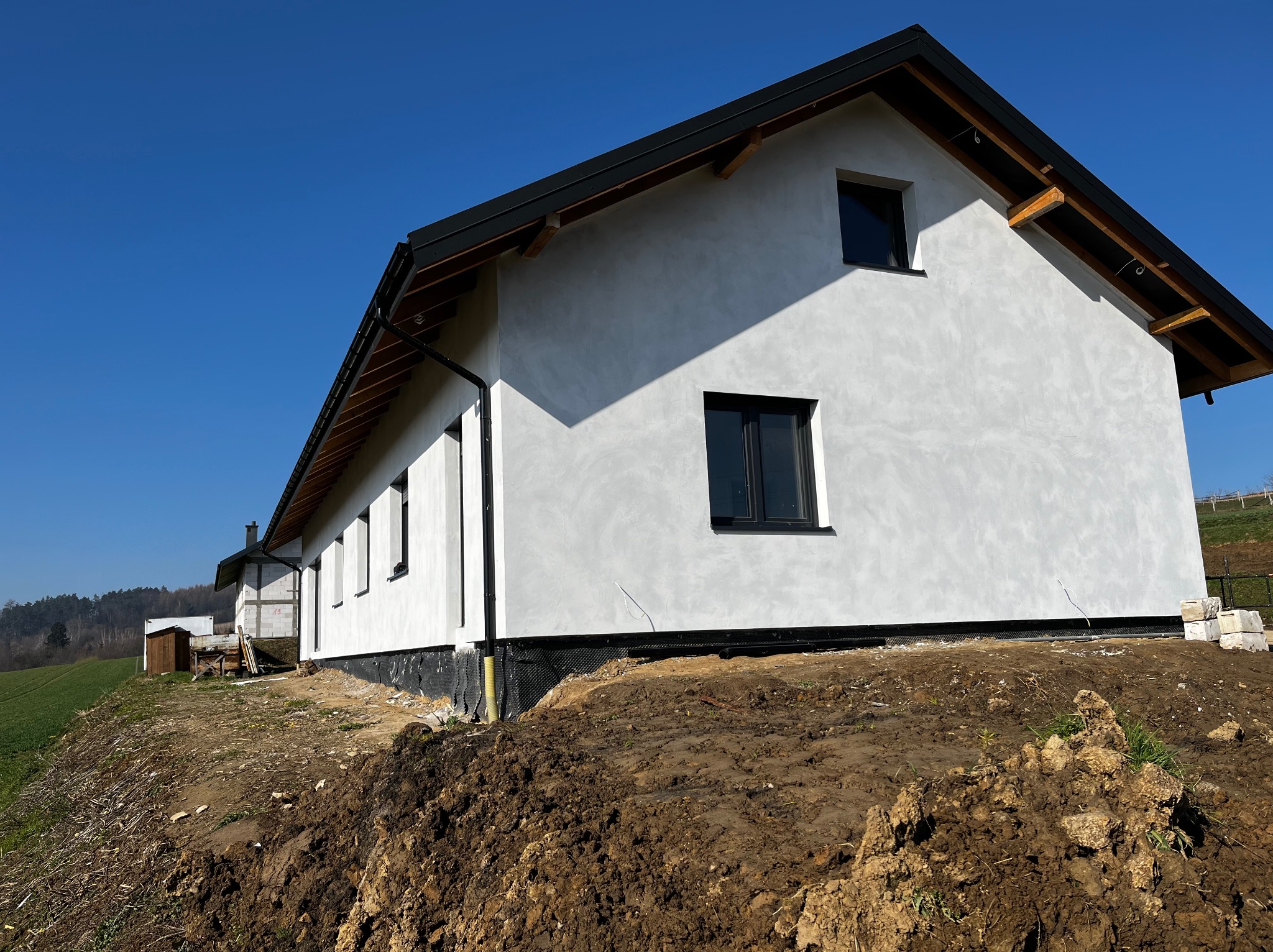 Surowa elewacja domu jednorodzinnego w trakcie prac wykończeniowych, widoczne czarne okna i fragment dachu, teren wokół domu jeszcze niezagospodarowany, świeżo usypana ziemia.