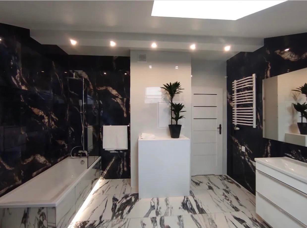 Nowoczesna łazienka z czarnymi marmurowymi płytkami na ścianach, białą wanną z podświetleniem LED, białą umywalką i białą szafką, białą grzejnikową suszarką oraz rośliną doniczkową na białym...