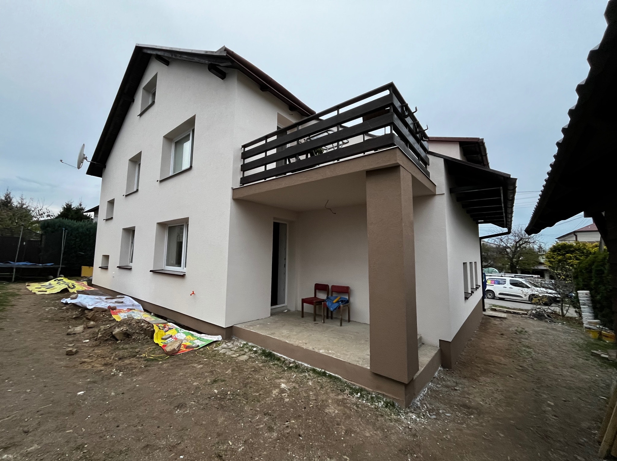 Świeżo wykończona elewacja domu jednorodzinnego z brązowymi akcentami, widoczne zabezpieczenia na ziemi i dwa krzesła na tarasie.